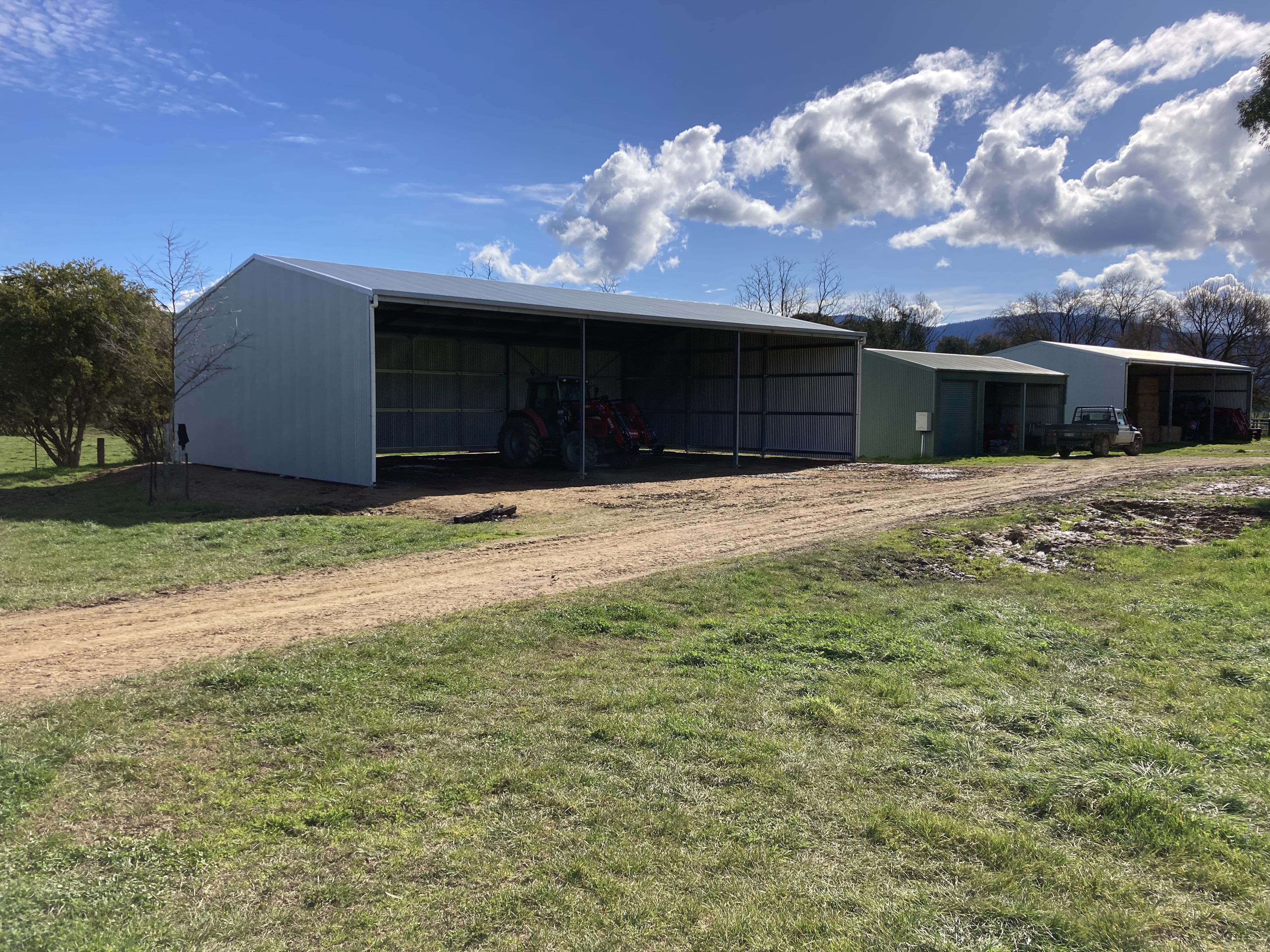 Open metal shed with a tractor inside, green grass field, dirt road, blue sky, and clouds.