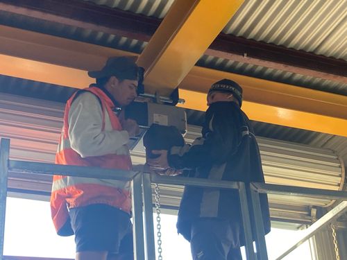 Two workers in safety gear inspecting or repairing equipment on a yellow overhead beam.