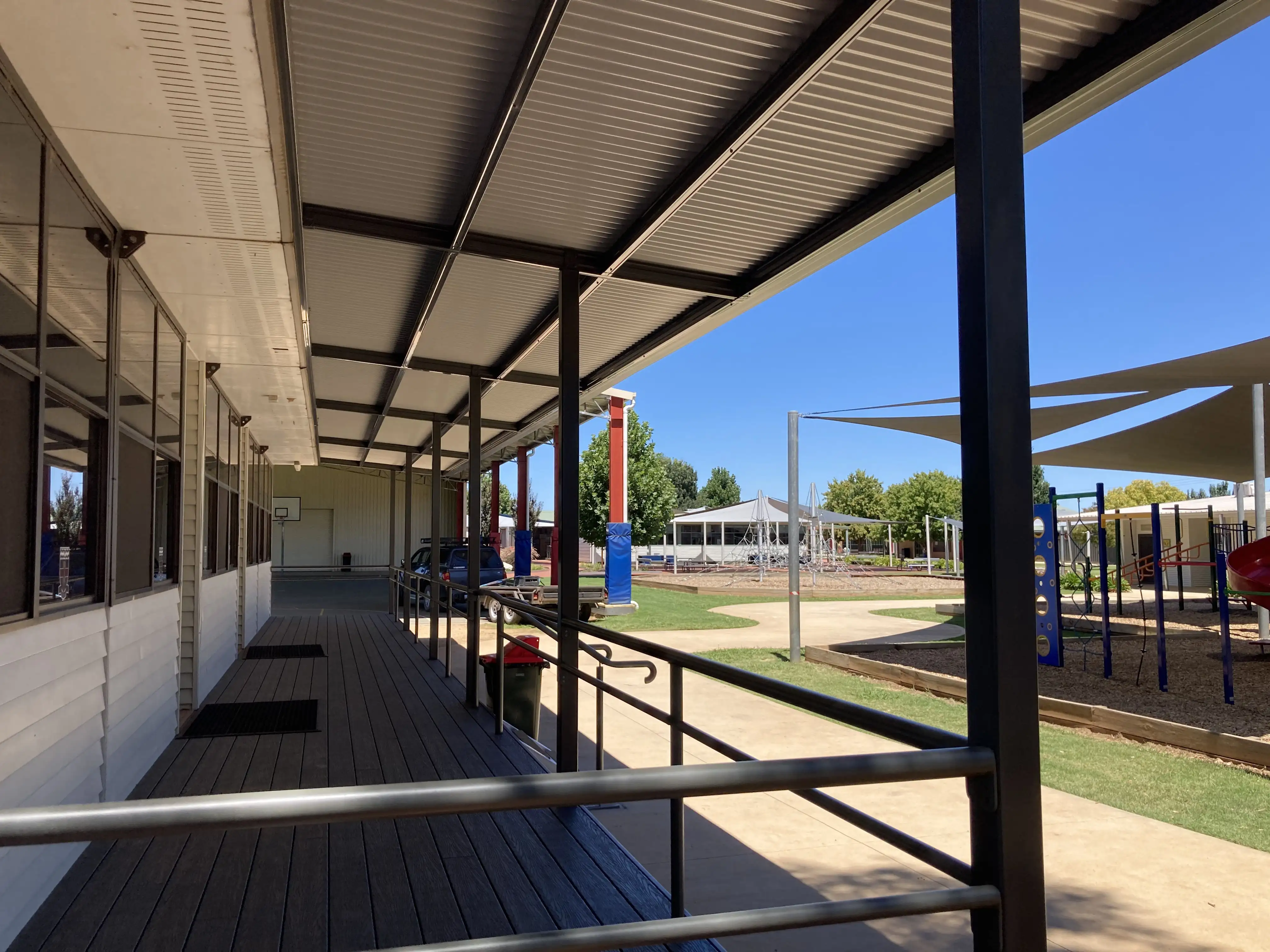 Covered walkway with railings beside a building overlooking an outdoor playground with shade sails.
