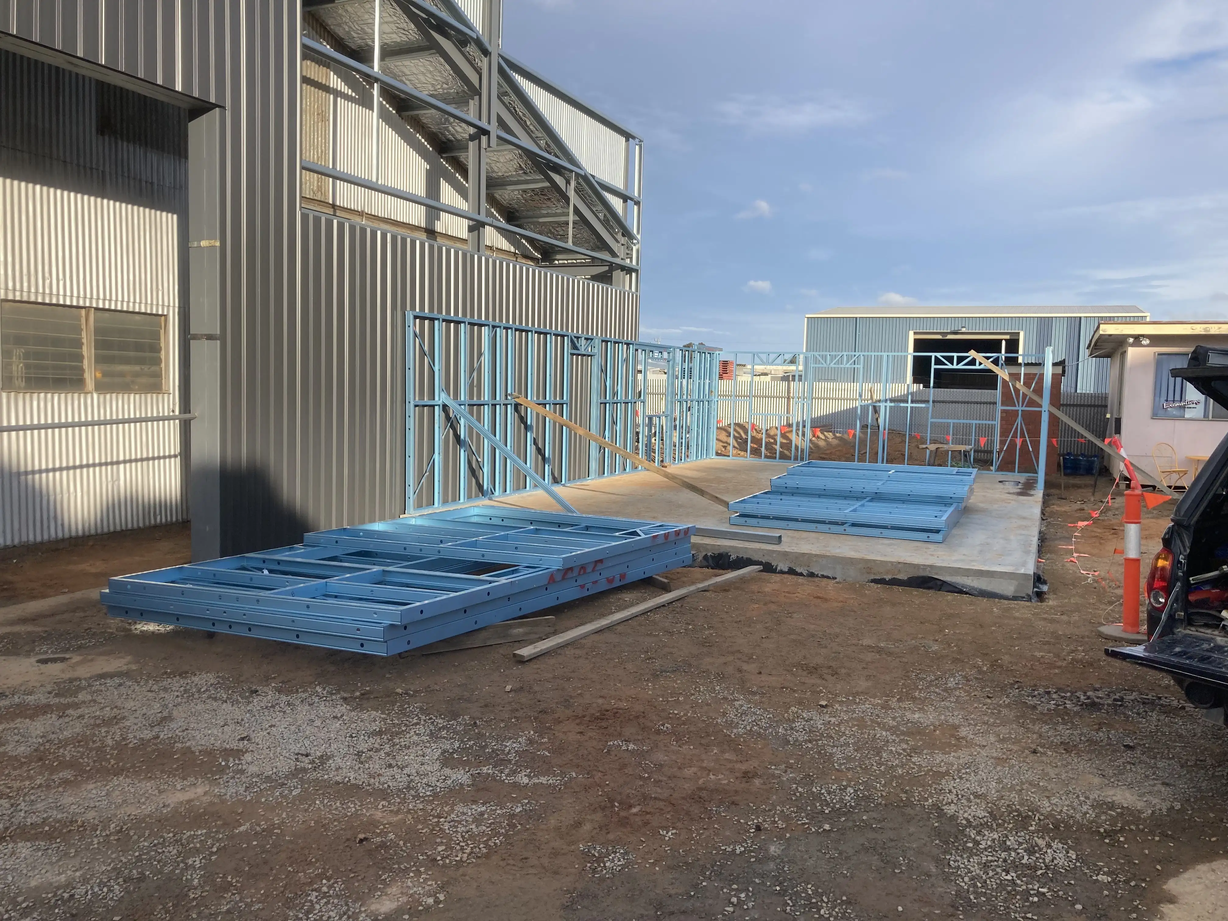Construction site with blue metal frames stacked and partially assembled on a concrete slab outdoors.