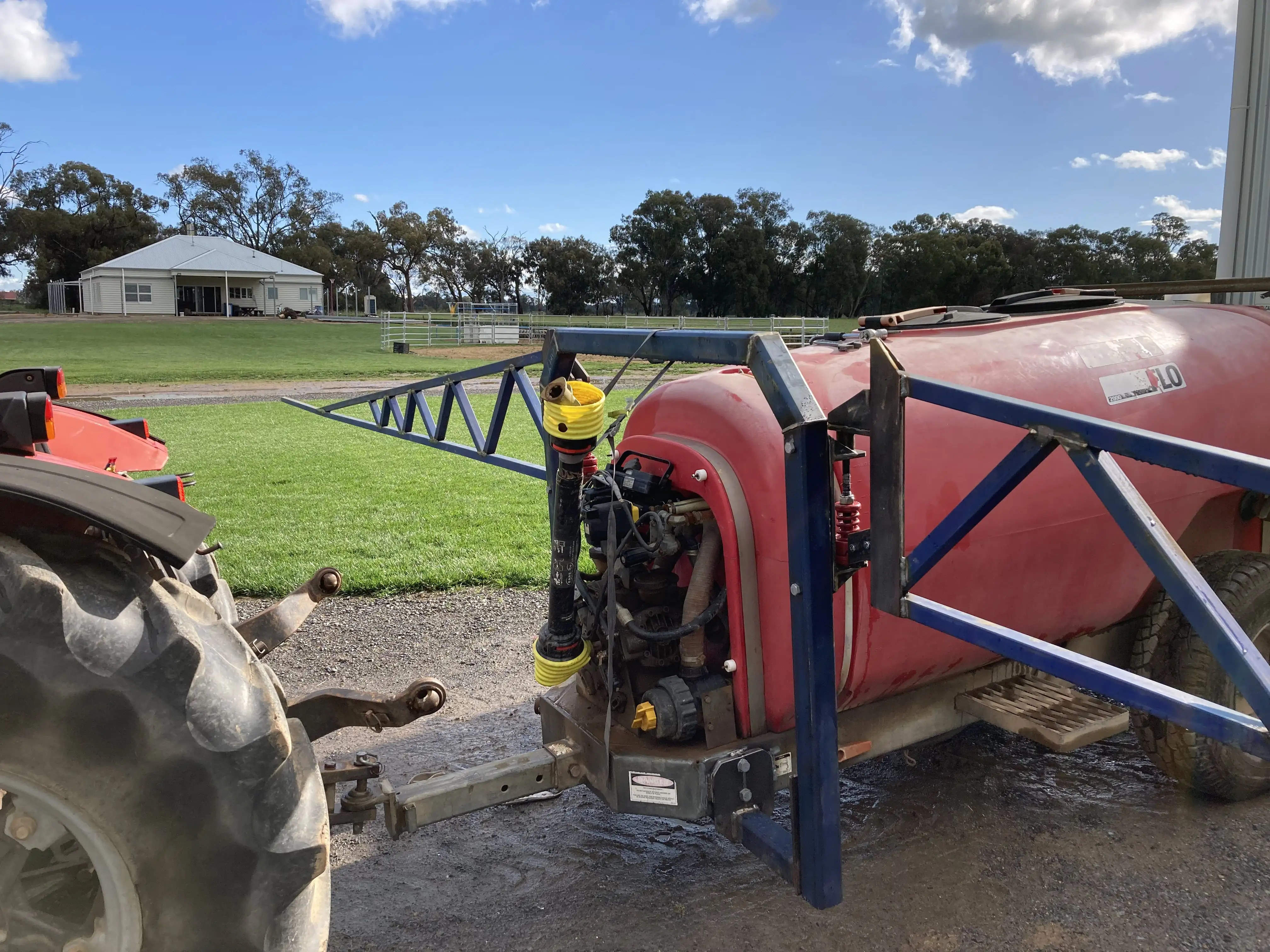 Red agricultural sprayer attached to tractor wheel, with green field and building in background.