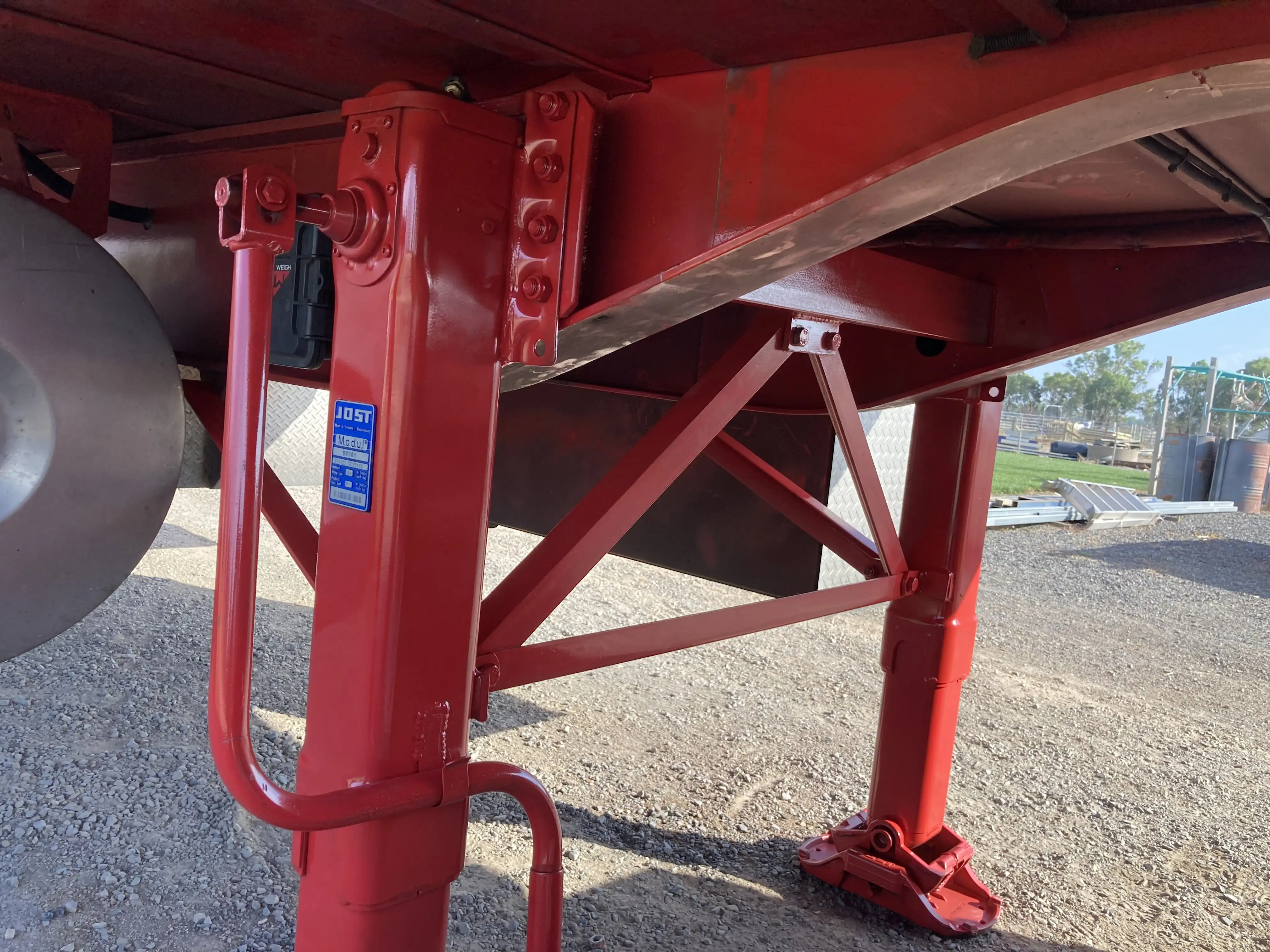 Close-up of red metal support legs and structural beams under a trailer on a gravel surface.
