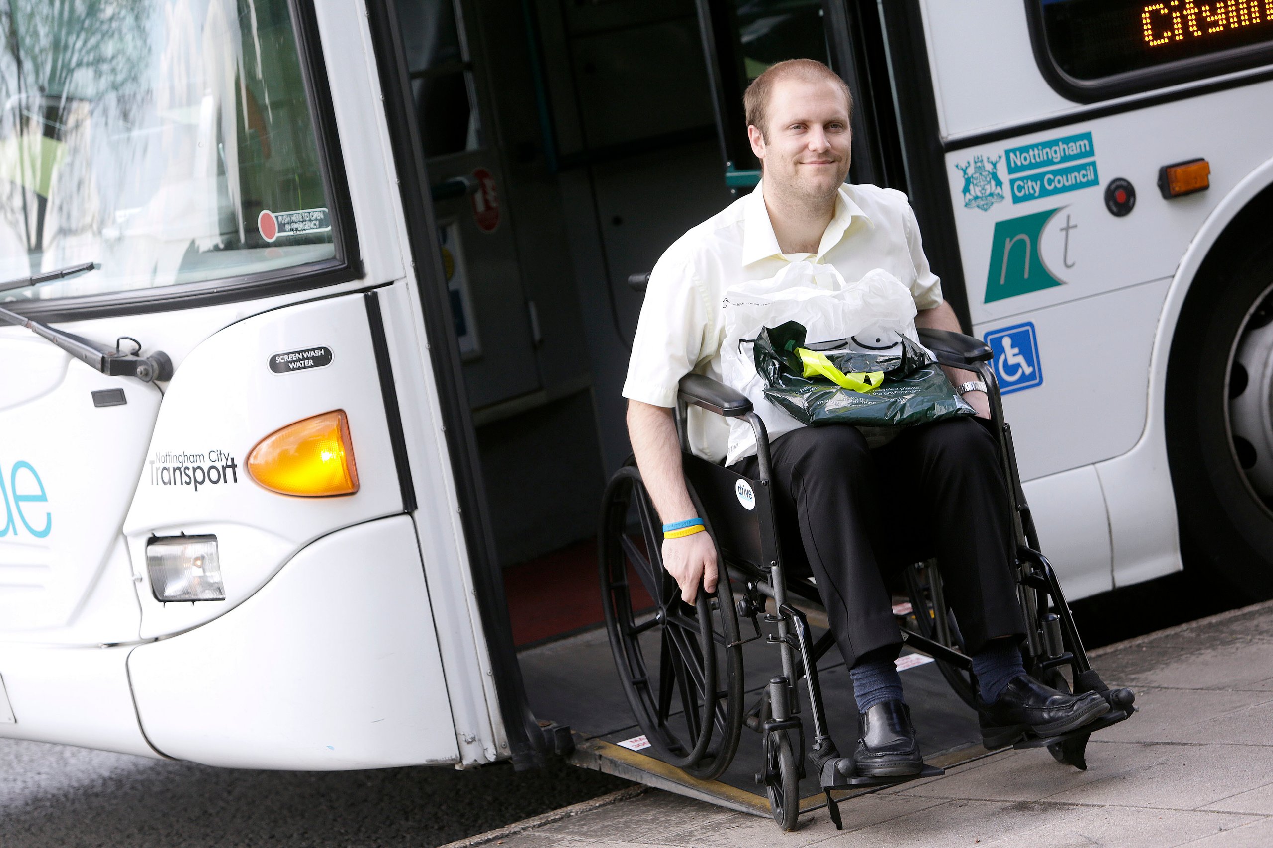 Man in wheelchair exiting a Nottingham City Transport bus via ramp, holding shopping bags in his lap.