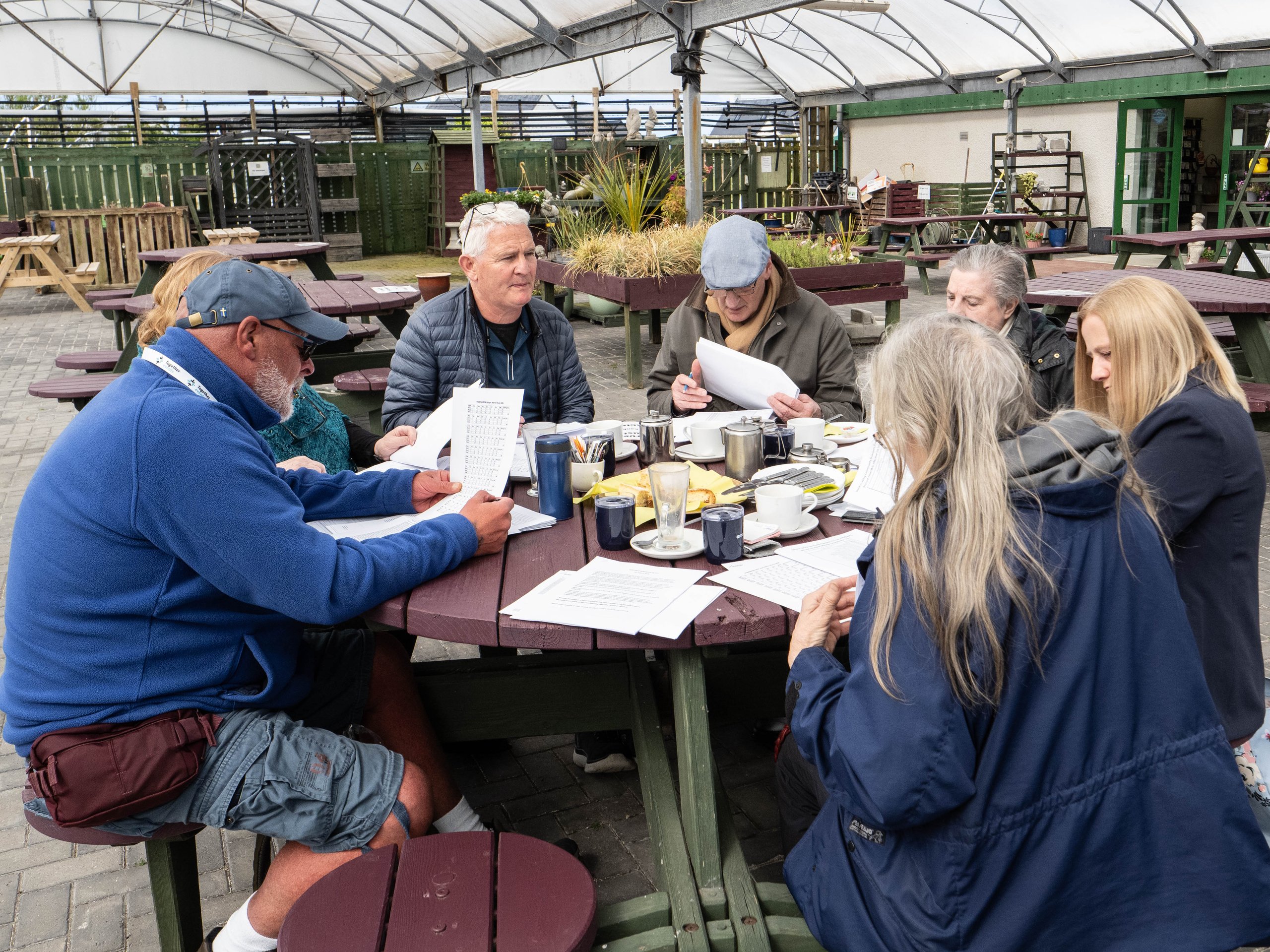 Group of elderly people sitting around a picnic table reading papers and drinking coffee outdoors.