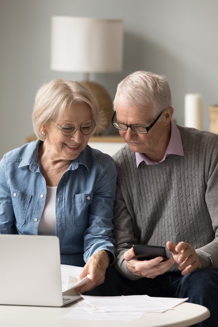 Smiling elderly couple with glasses reviewing papers and using a laptop and smartphone at a table.