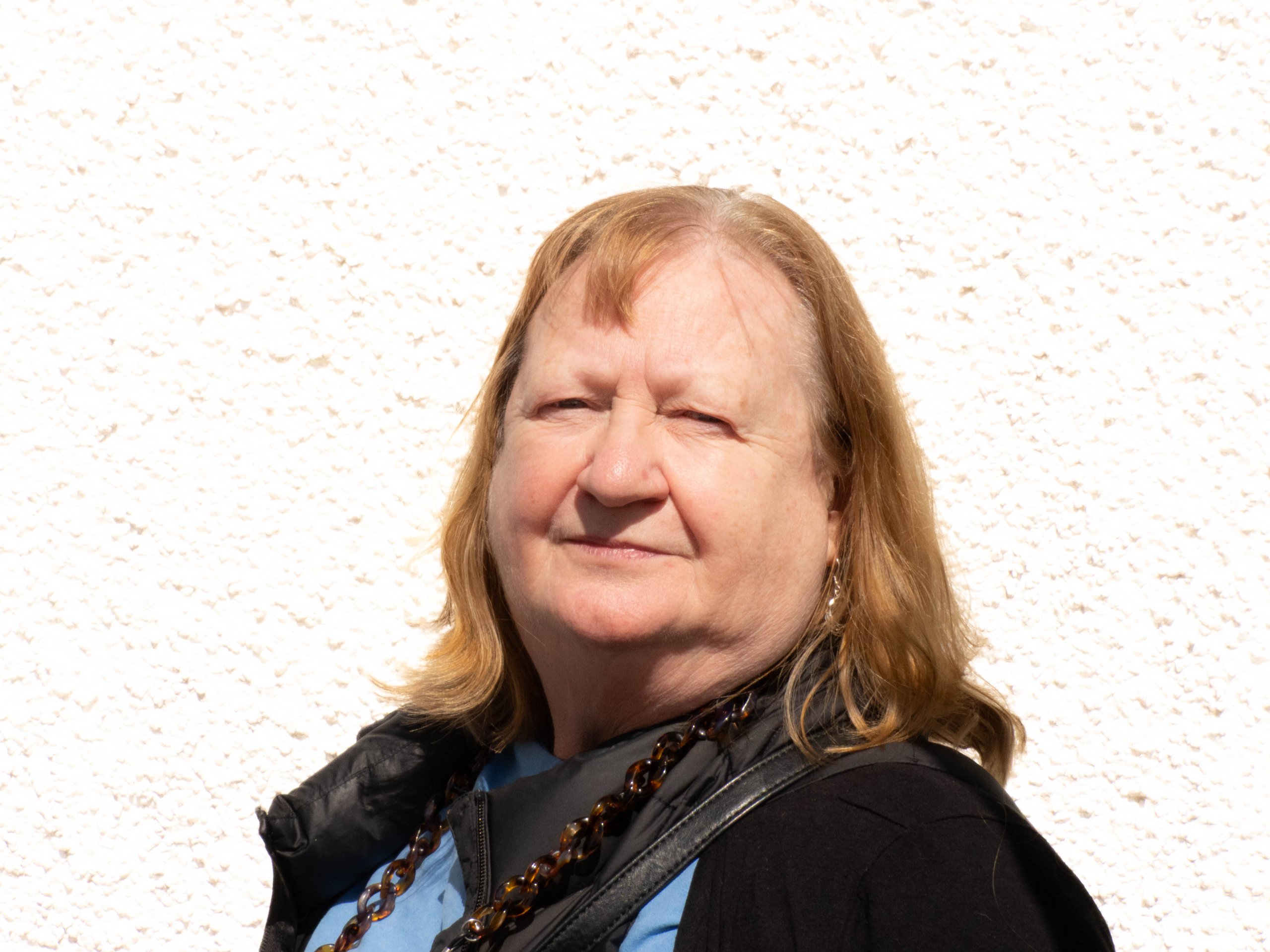 Older woman with light brown hair wearing a blue shirt, black jacket, and beaded necklace against a textured wall.