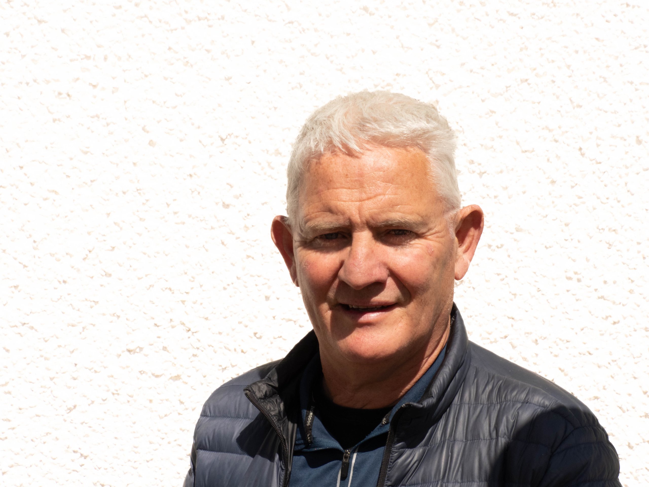 Older man with short white hair wearing a dark jacket and standing against a textured white wall.