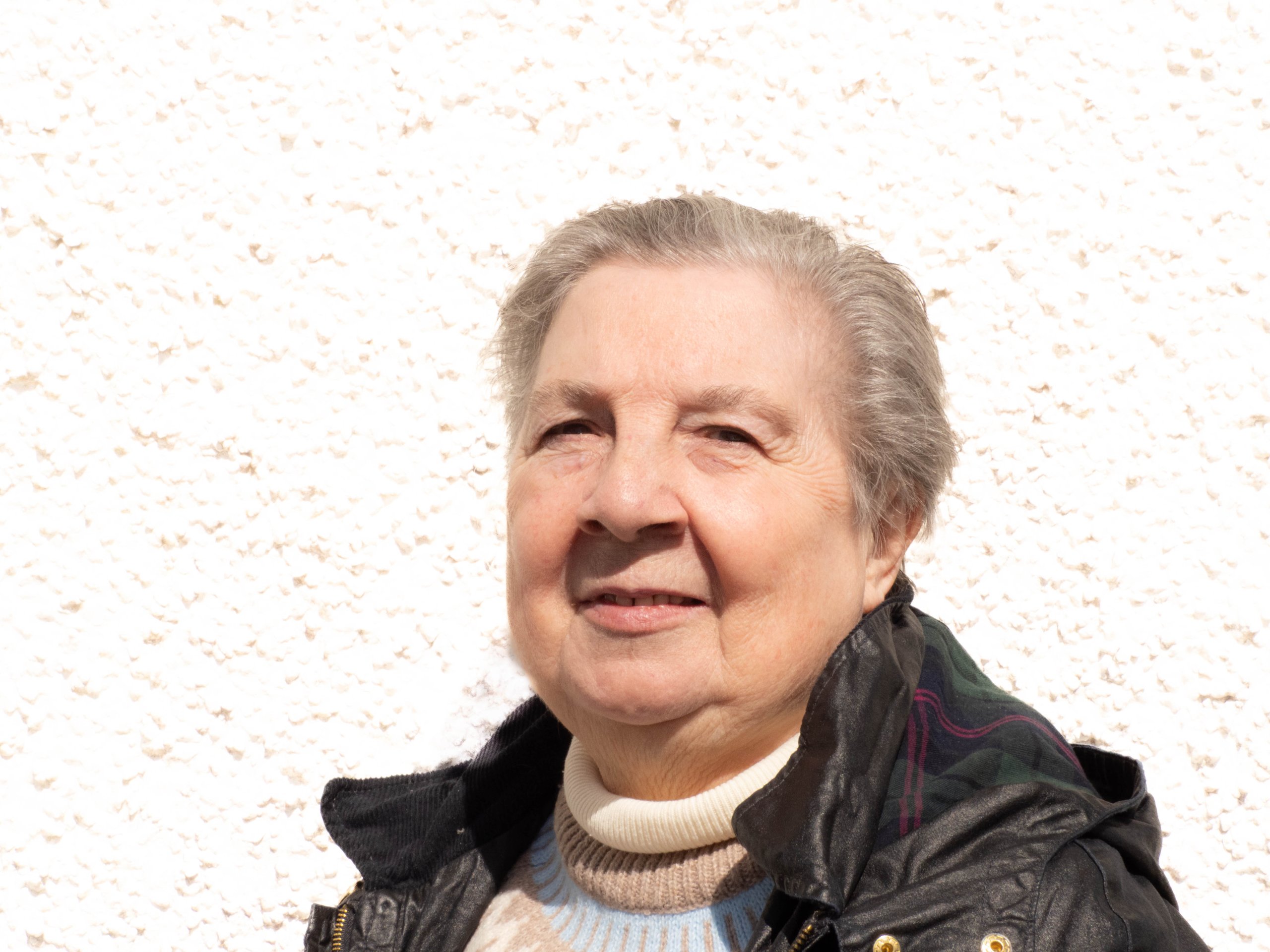 Portrait of an elderly woman with short gray hair wearing a jacket and sweater against a white wall.