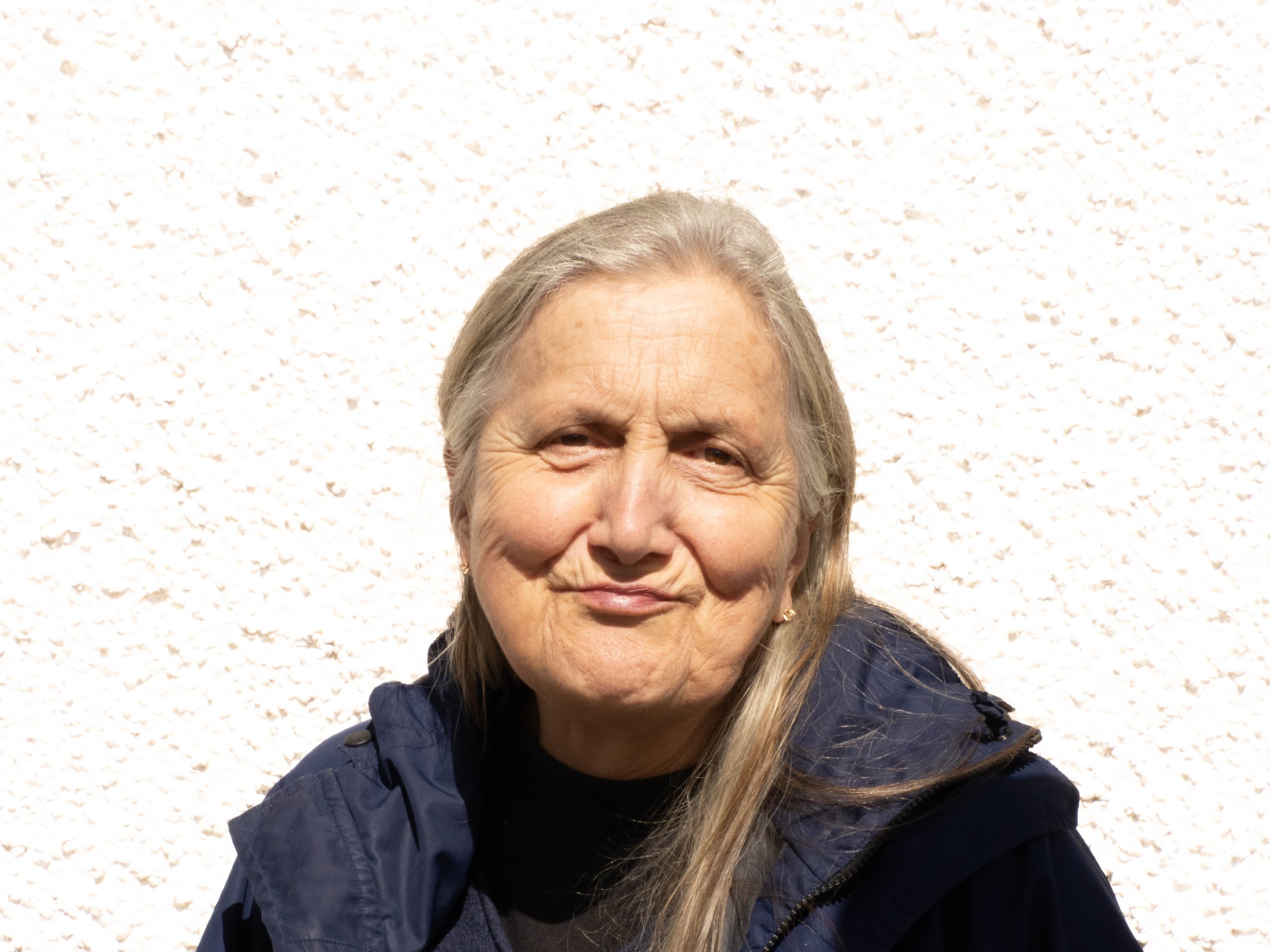 Older woman with long gray hair in a navy jacket standing against a light textured wall.