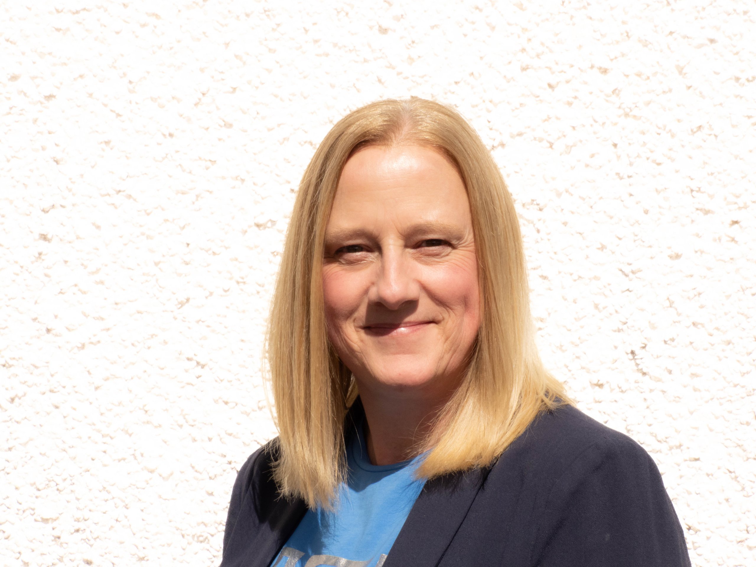 Smiling woman with straight blonde hair wearing a blue top and dark blazer against a white textured wall.