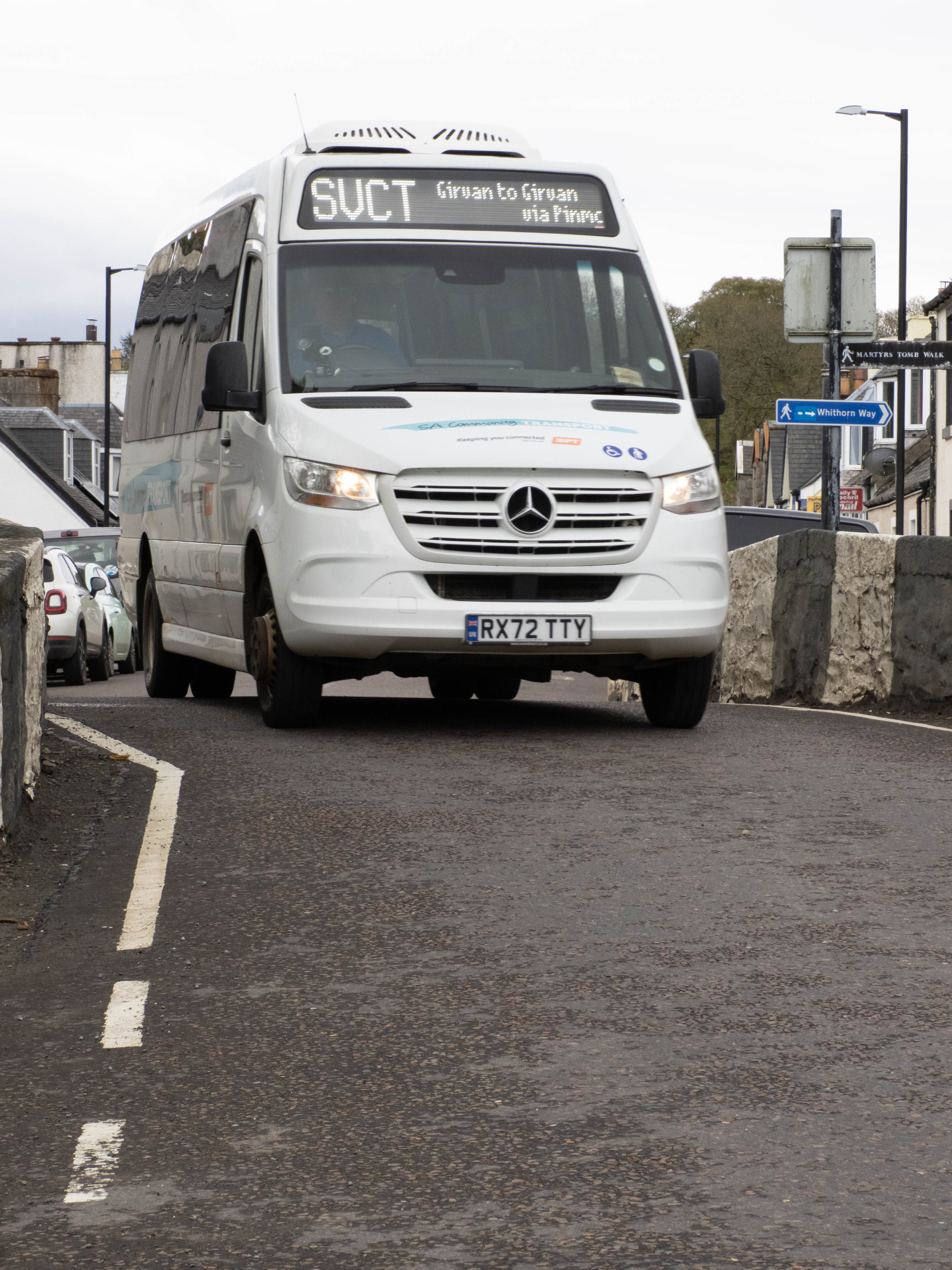 White Mercedes van with license RX72 TTY driving on a road with stone walls and street signs nearby.