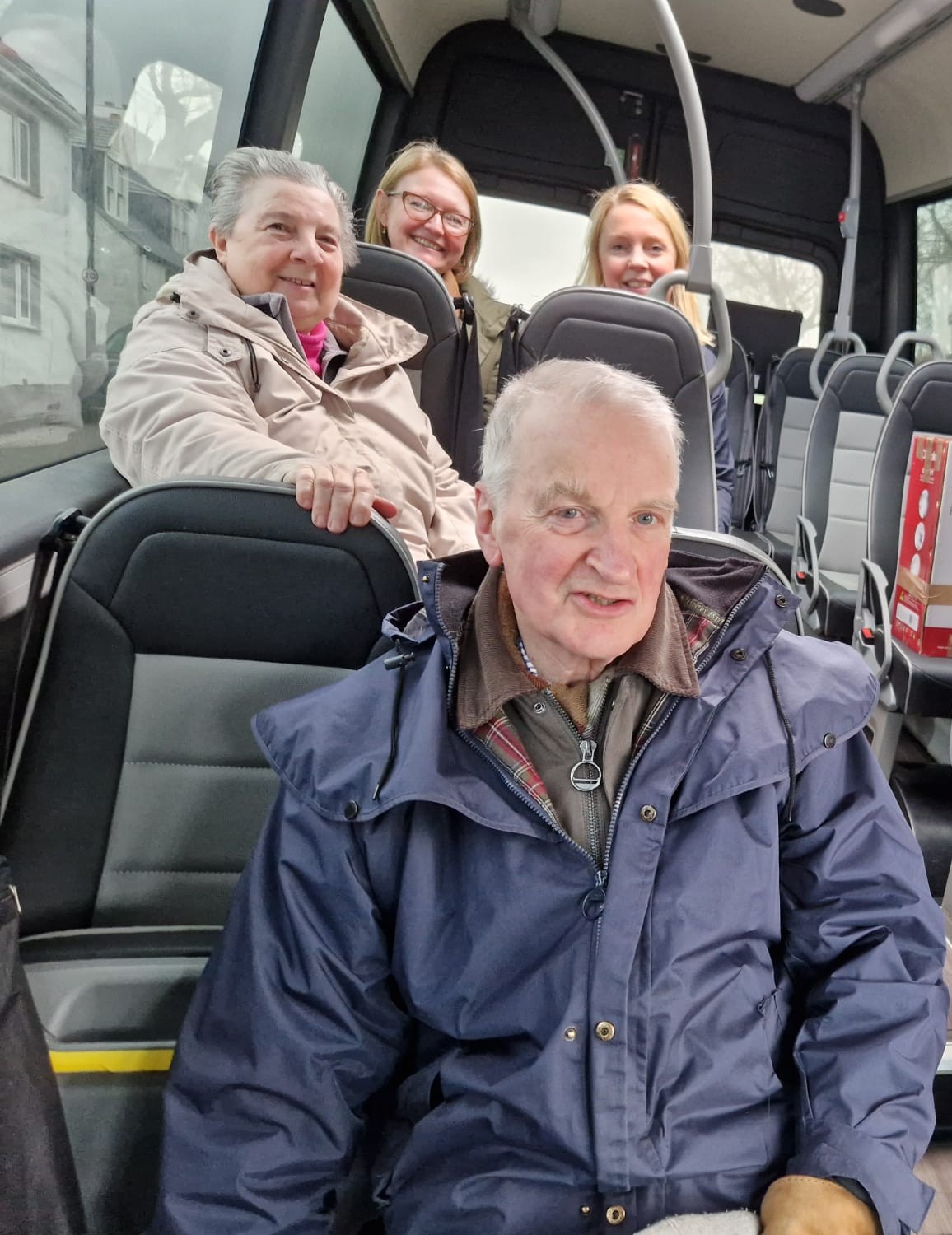 Four people seated inside a bus, smiling toward the camera, with empty seats visible behind them.
