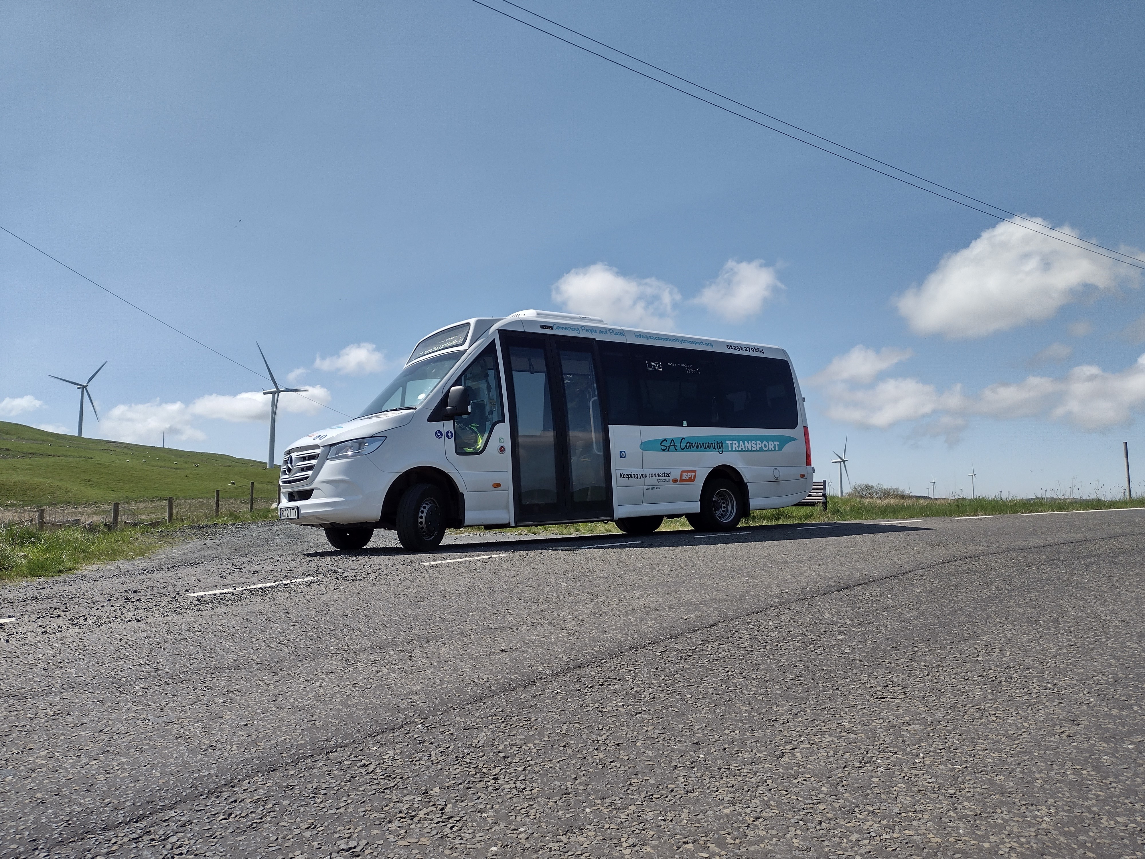 White community transport minibus parked on a rural road with wind turbines on grassy hills.