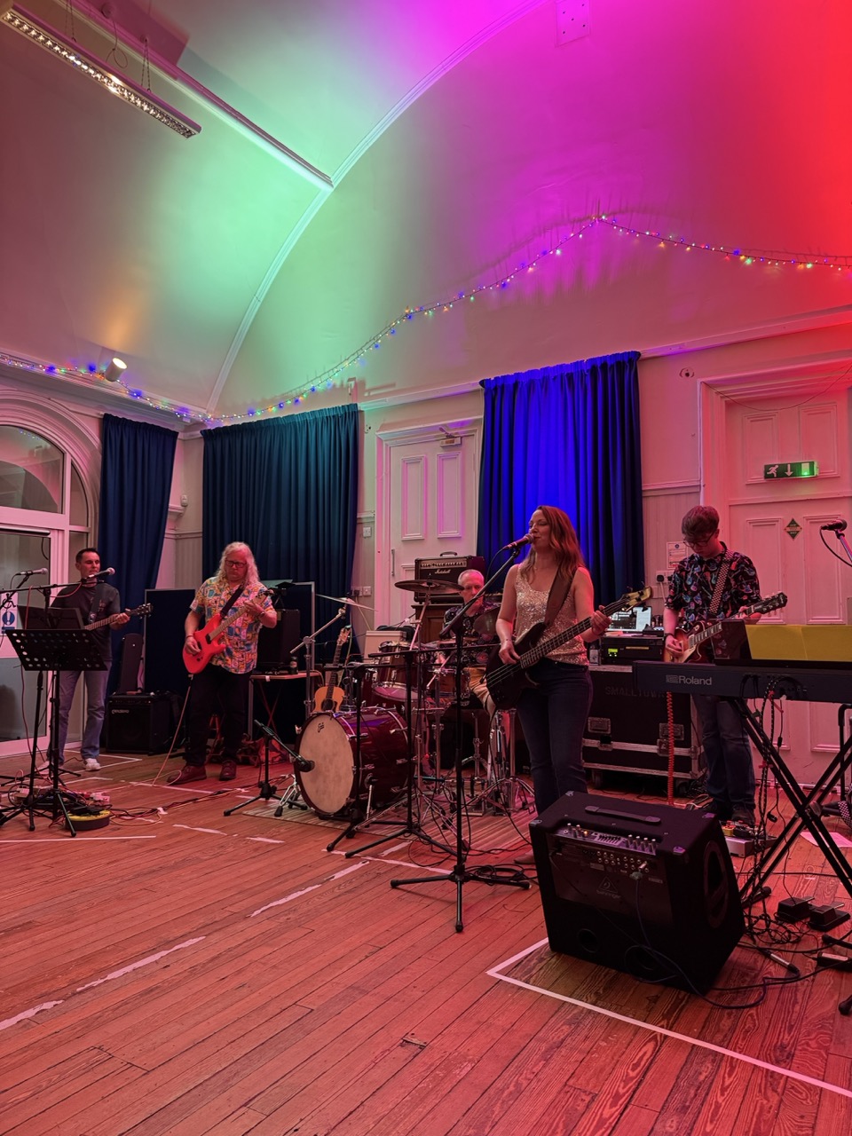 Five musicians performing on stage with colored lights and musical instruments in a wooden-floor room.