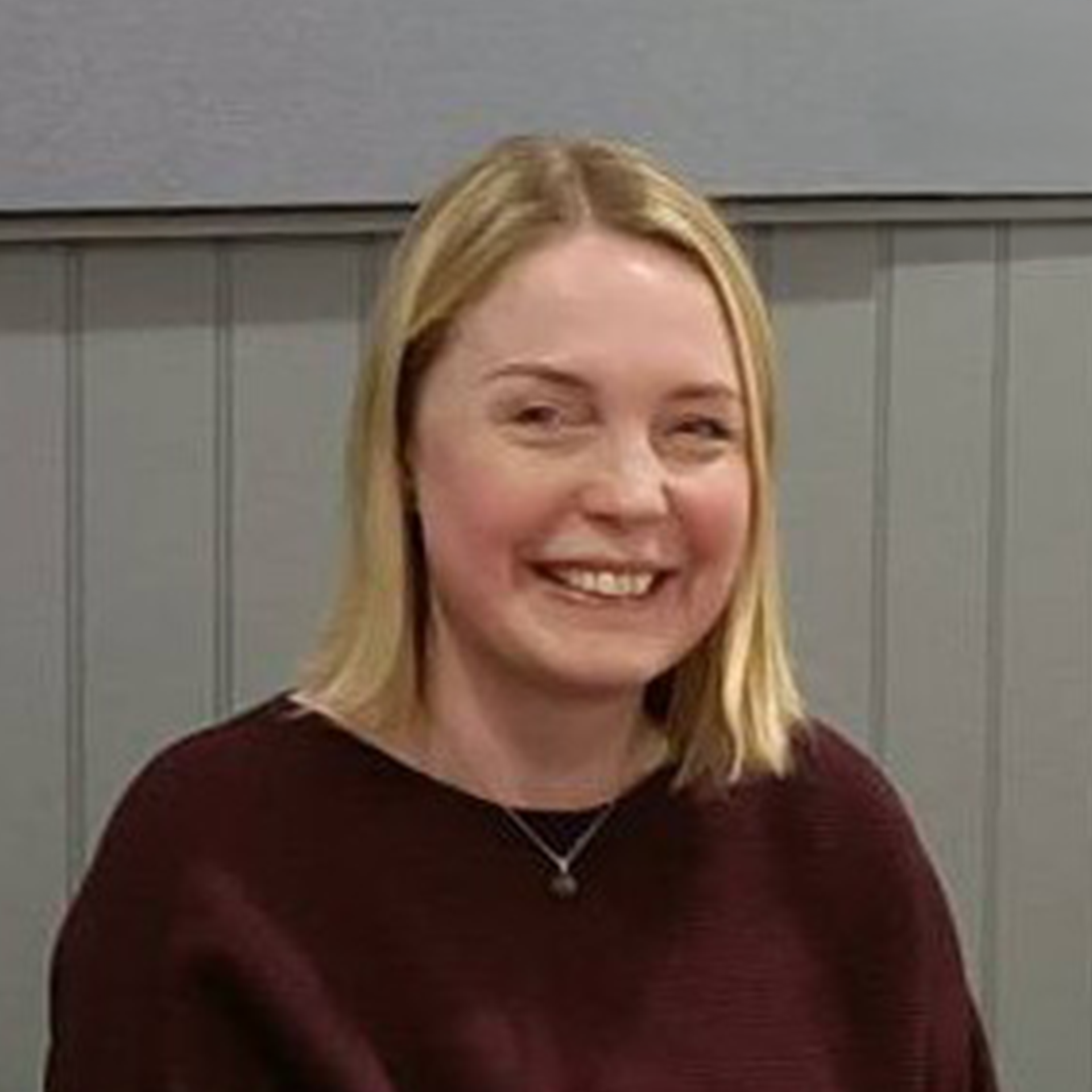 Smiling woman with straight blonde hair wearing a blue top and dark blazer against a white textured wall.