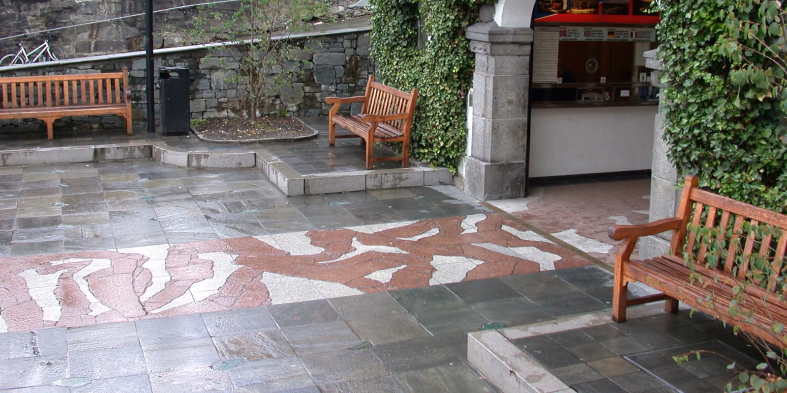 Outdoor mosaic pavement between wooden benches under ivy-covered stone arches.