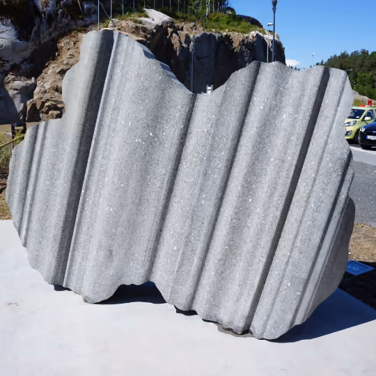 Grå betongskulptur med bølgete vertikale riller plassert utendørs på betongplattform.
