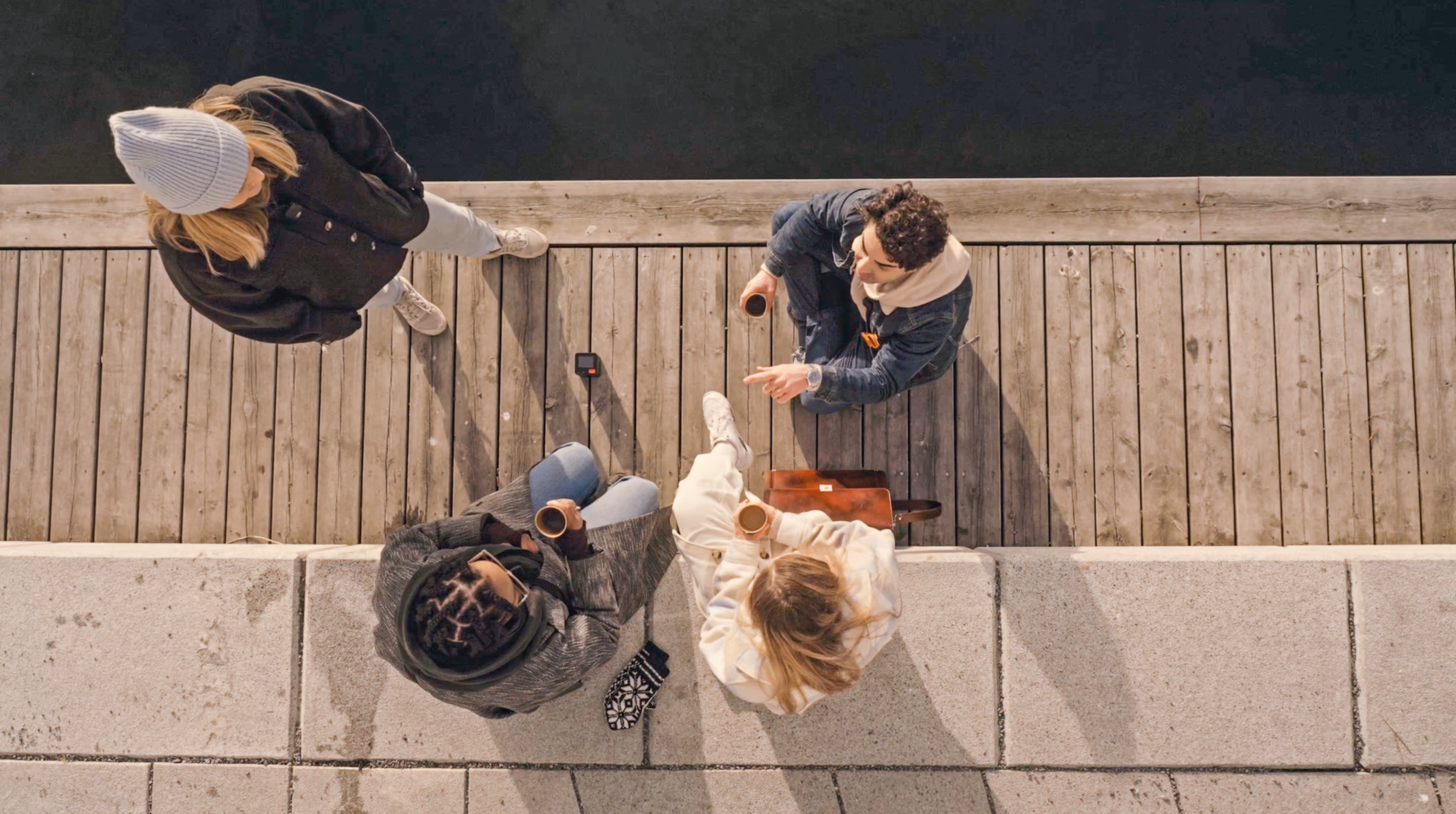 Aerial view of four people talking on a wooden dock by the water, two seated with coffee cups and two standing