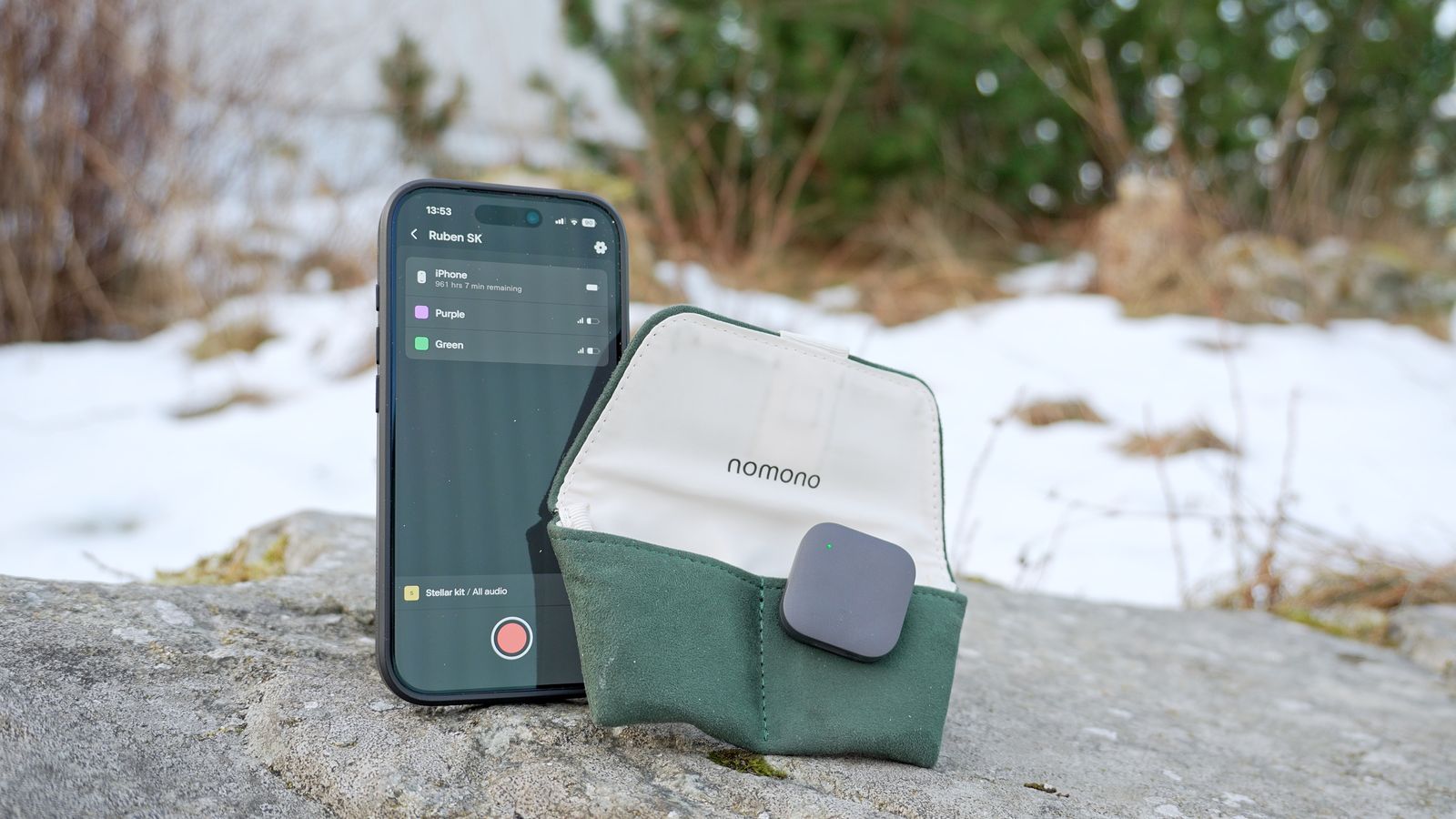 Smartphone running the Nomono recording app with two mics connected, propped beside an open green Nomono pouch holding a Stellar Mic on a rock in the snow.
