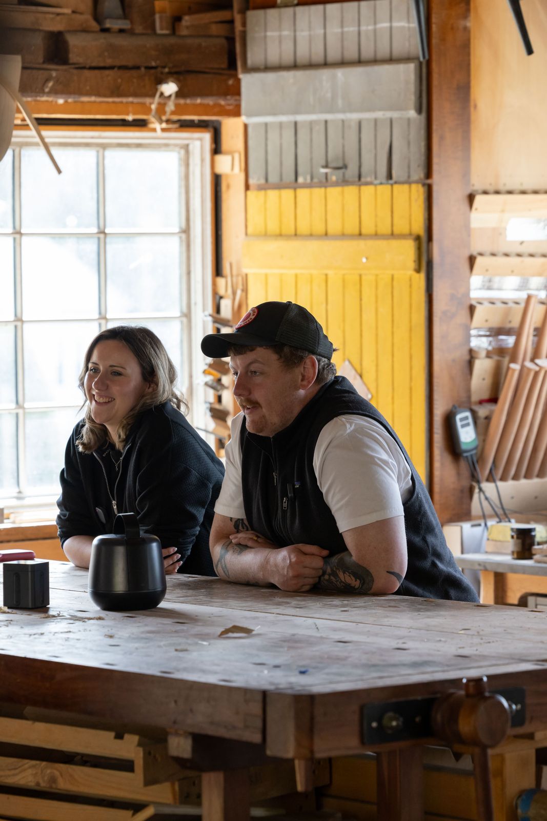 Two people leaning on a large workbench in a woodworking shop mid-conversation, with the Sound Capsule case and Space Recorder on the bench.