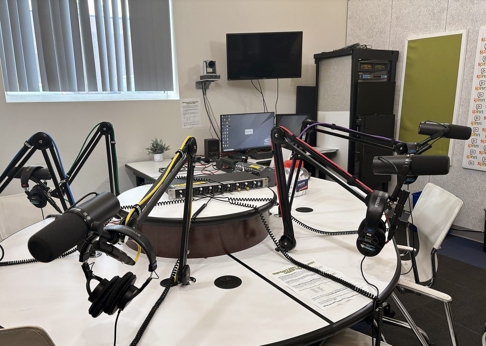 Round podcast table with several Shure SM7-style dynamic microphones on colour-coded RØDE boom arms, studio headphones and an audio interface rack in the middle