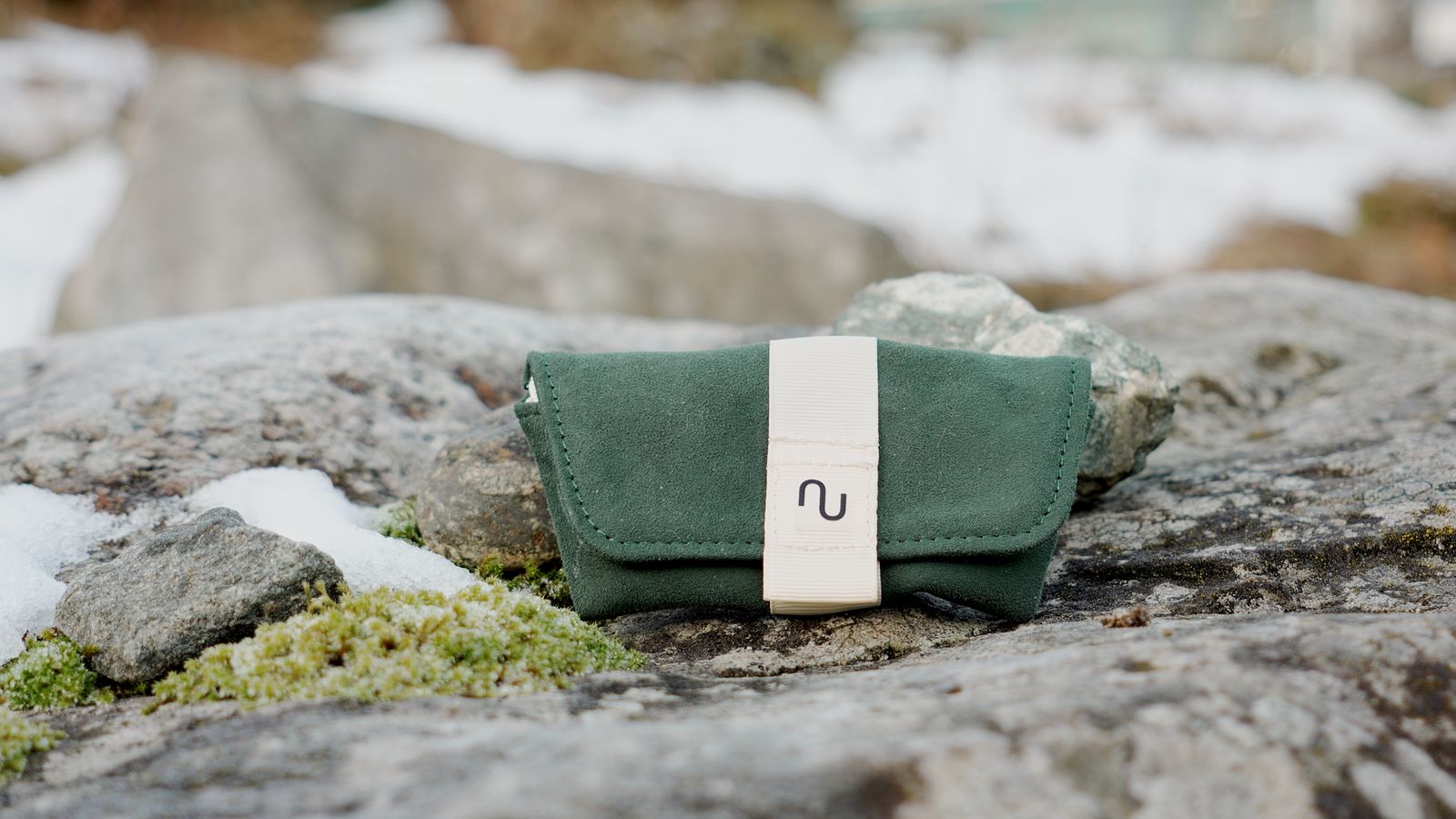 Closed green suede Nomono carry pouch with a cream strap resting on a mossy rock with patches of snow.