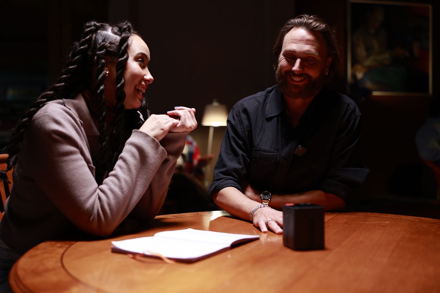 Woman and man laughing at a round wooden table with an open notebook and the Space Recorder between them; a small clip-on mic is visible on his shirt.