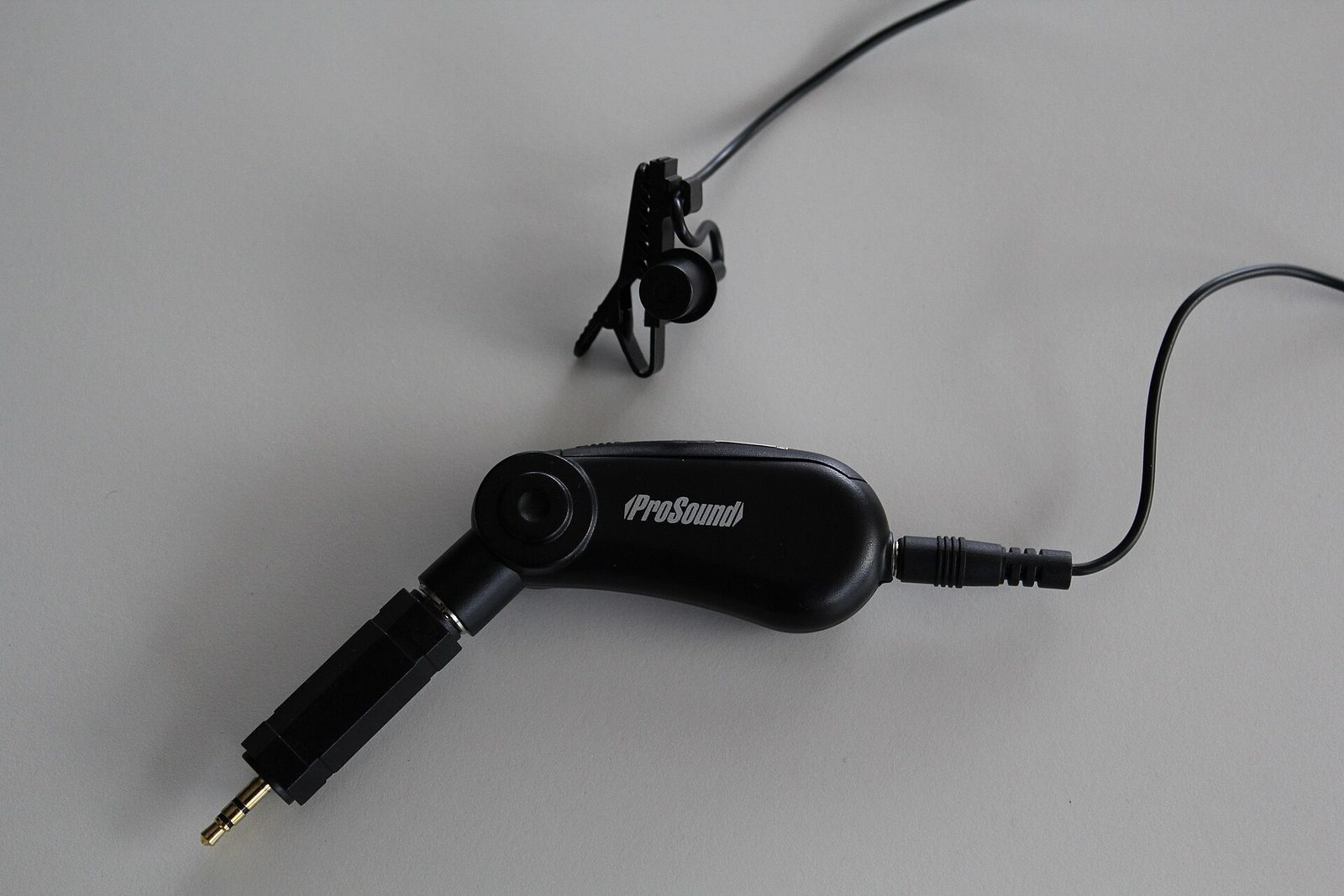 Black clip-on lavalier microphone with its tie clip, cable and battery module ending in a 3.5 mm jack, on a grey surface