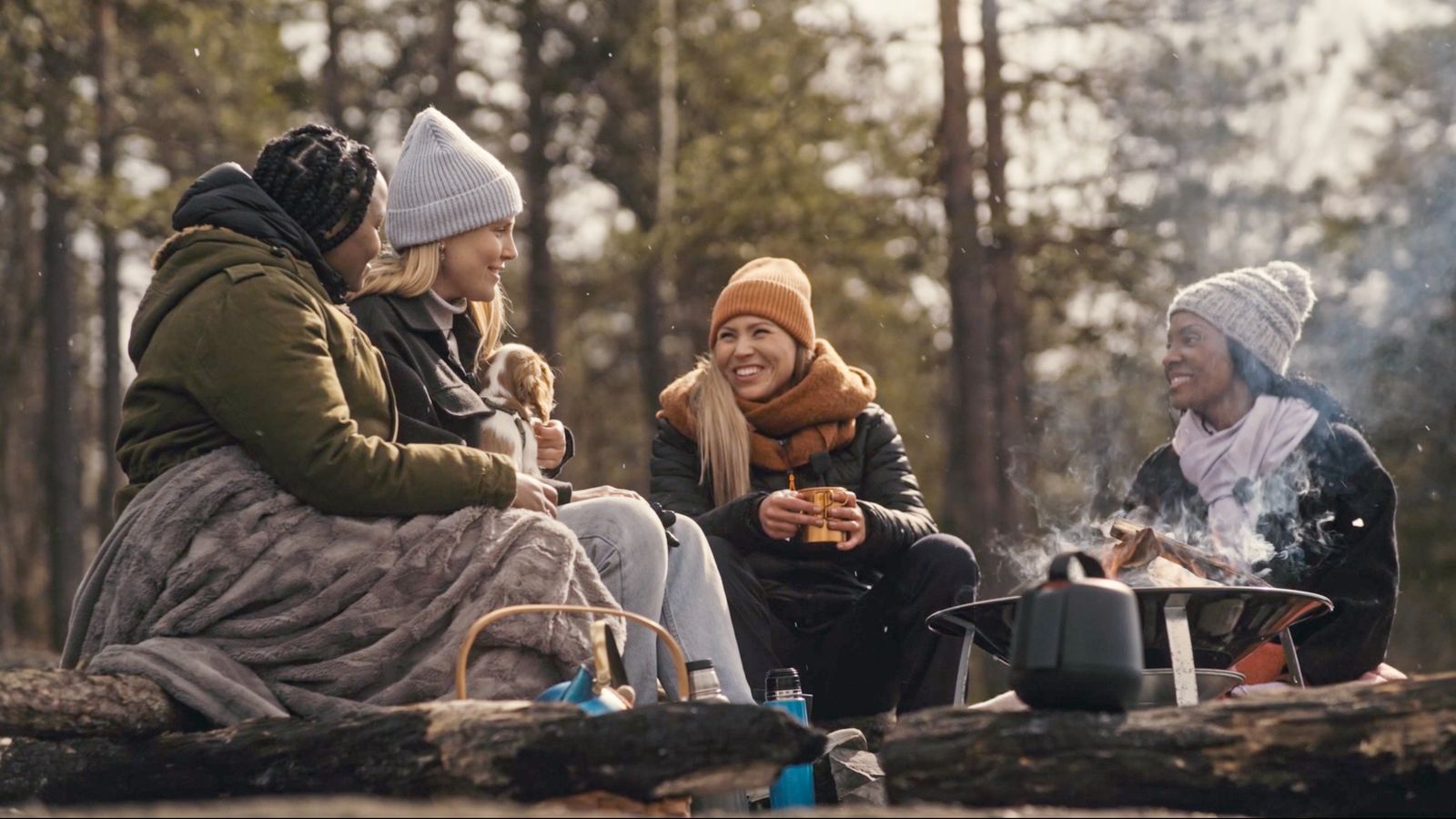 Four women in winter clothes chatting around a fire pit in a forest, with the Sound Capsule case on a log in the foreground.