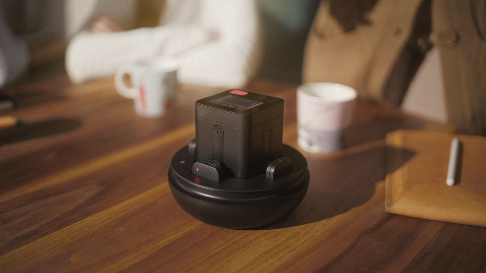 Space Recorder on its charging base with docked mics on a wooden cafe table, mugs and people blurred in the background.