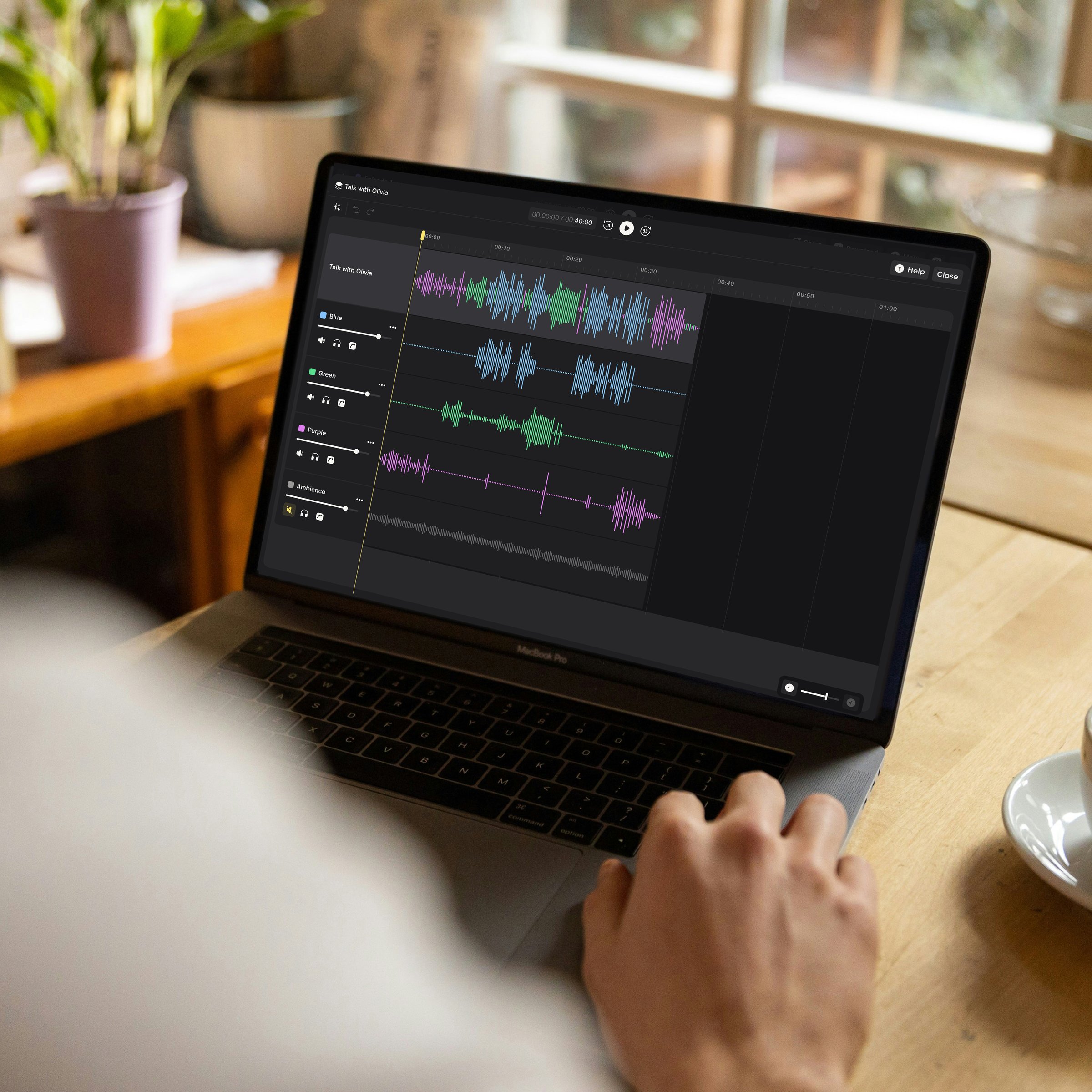 Person at a wooden café table editing a recording in Nomono Studio Cloud on a laptop, with speaker tracks and an Ambience track on screen