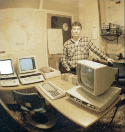 Pete Morris standing in his radio shack. Computers and a printer fill the desk in front of him, with a large monitor nearest the camera, a world map on the wall to his left and radio equipment on a shelf to his right