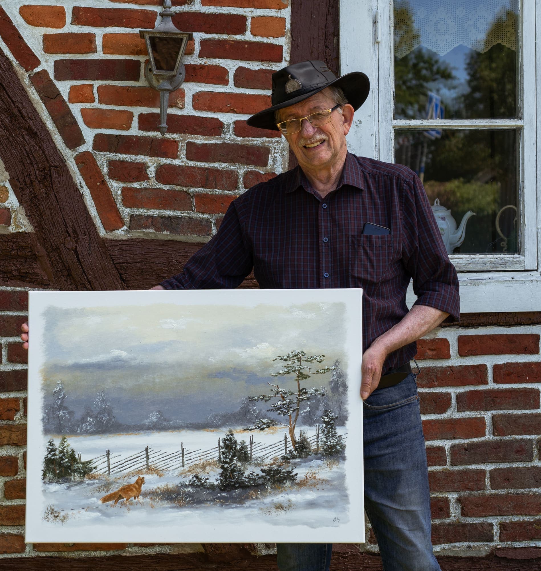 Konstnären Ingvar Stendahl framför sitt korsvirkeshus med en målning.