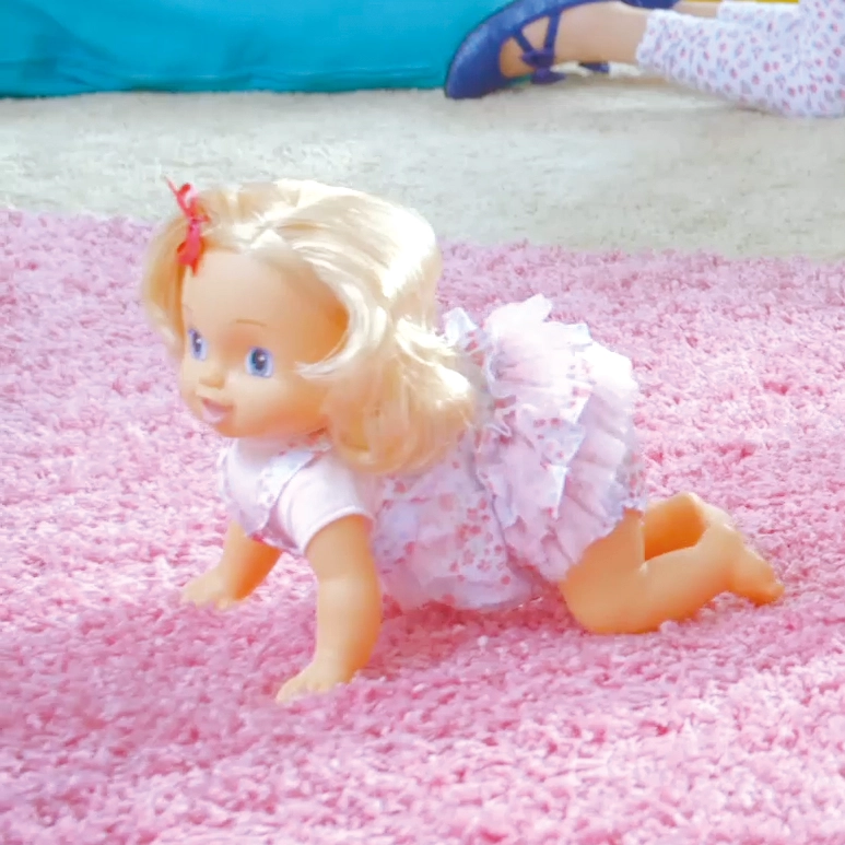 Blonde baby doll with a pink dress crawling on a pink carpet, child wearing blue shoes in background.
