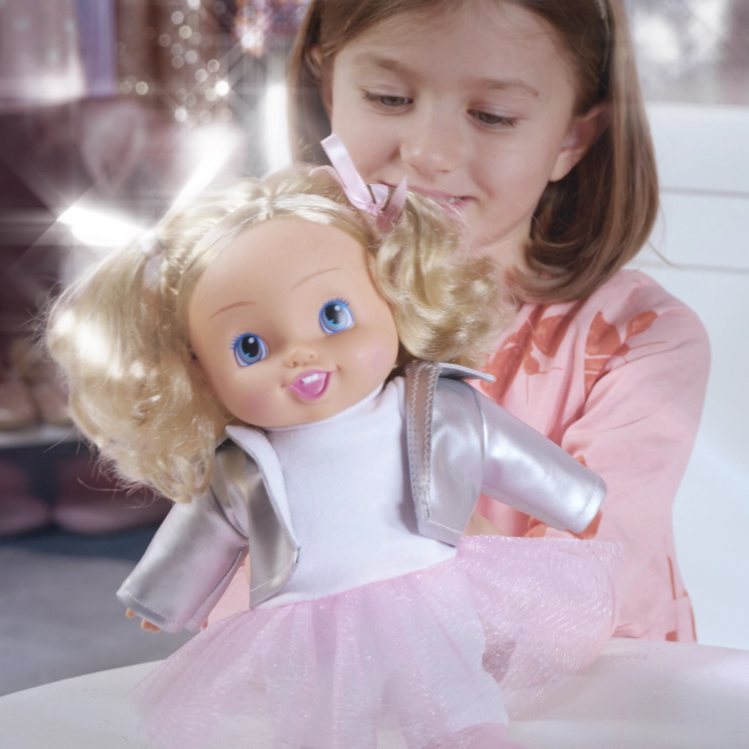 Smiling girl holding a doll with blonde hair, blue eyes, silver jacket, and pink tutu skirt.