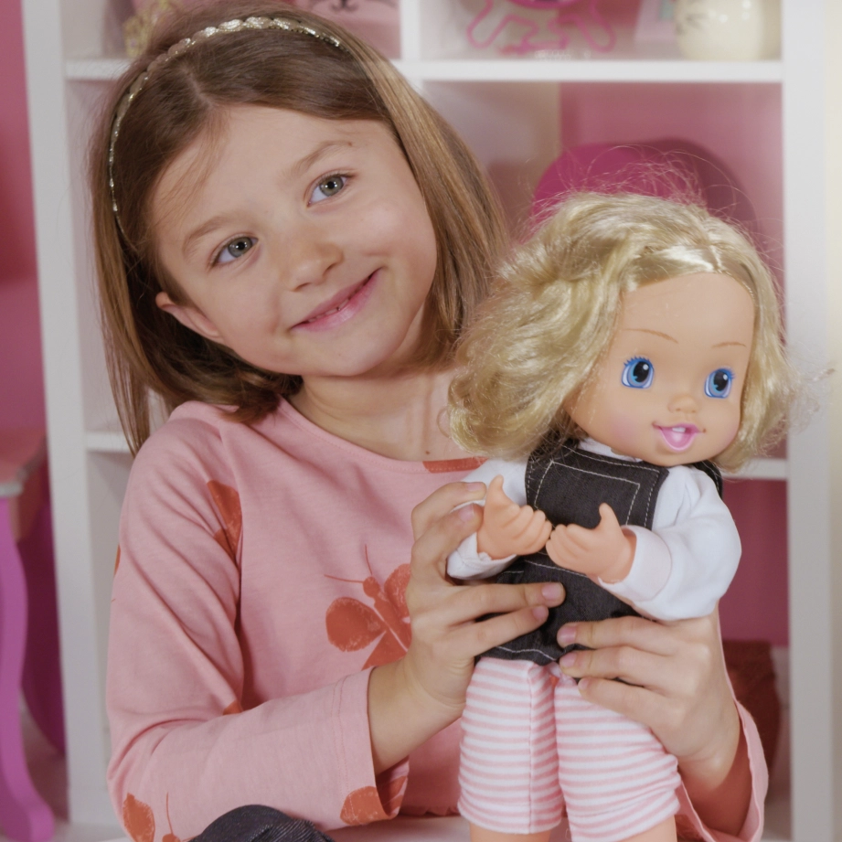 Smiling girl in pink shirt holding a blonde doll with blue eyes wearing striped pants.