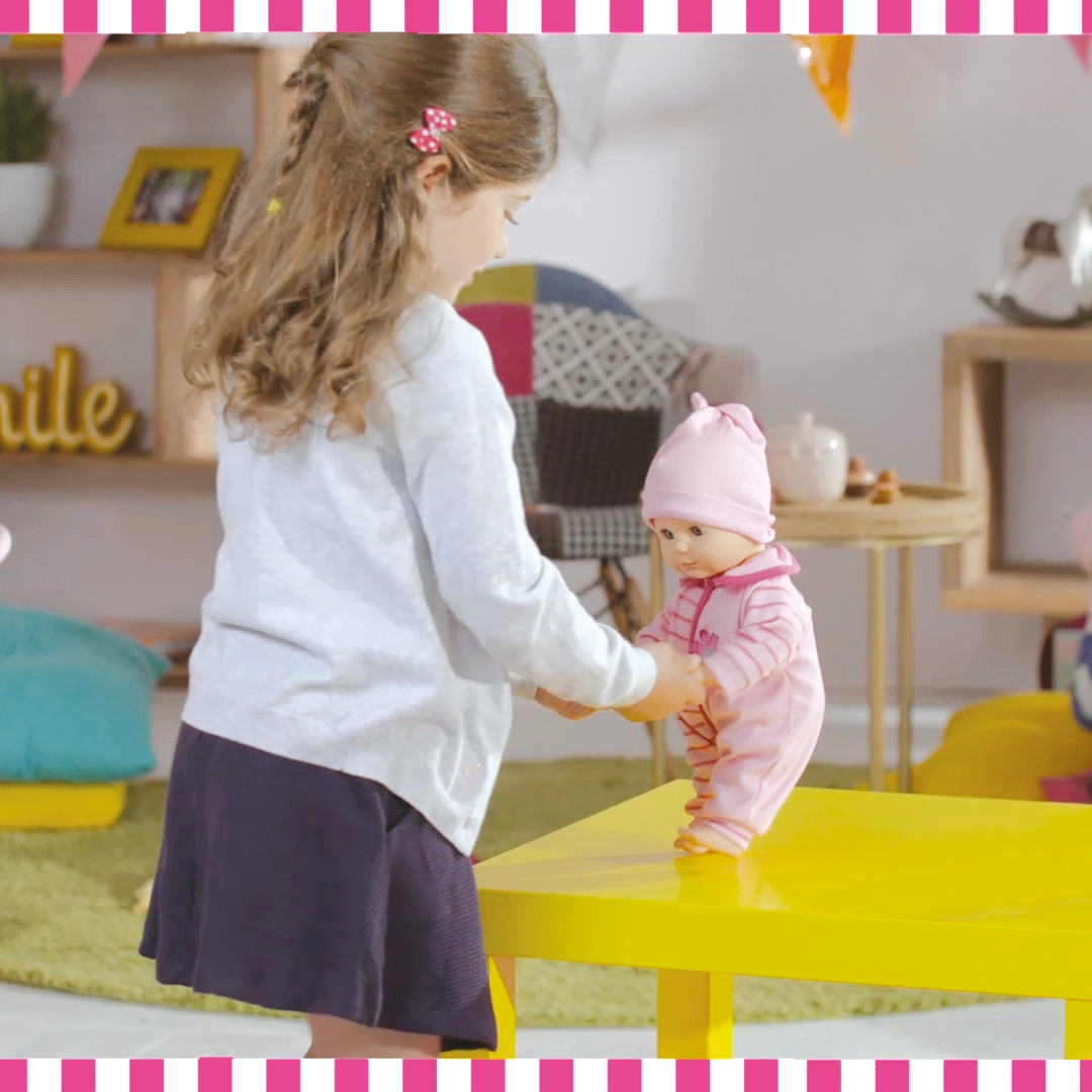 Little girl holding hands of a baby doll dressed in pink standing on a yellow table.