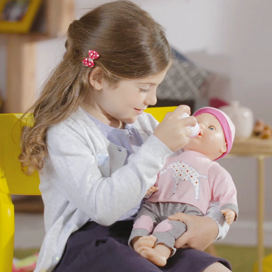 A girl with a red bow feeds a baby doll with a bottle while sitting on a yellow chair.
