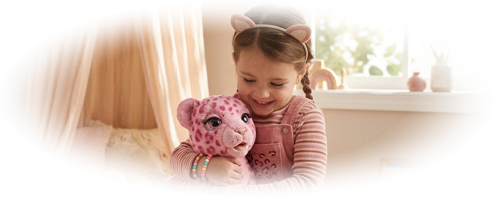 A smiling girl in pink dress hugs a pink spotted stuffed leopard while sitting on a bed.