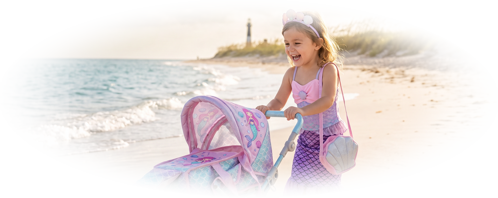 Young girl dressed as a mermaid pushing a matching mermaid-themed doll stroller on the beach.