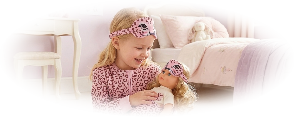 Girl in pink leopard pajamas and mask holding a doll with matching pajamas and mask on a furry rug.