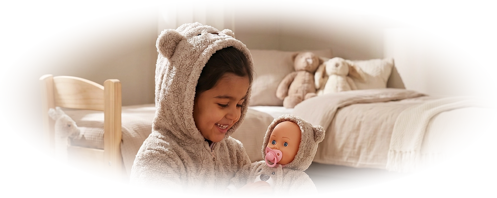Child in a bear onesie playing with a matching bear-themed doll in a cozy bedroom.