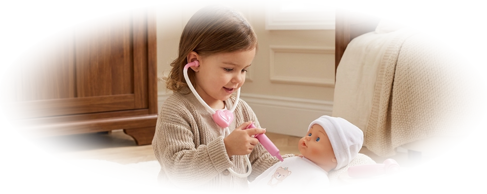 A girl in a beige sweater plays doctor using a toy stethoscope on a baby doll in white clothes.