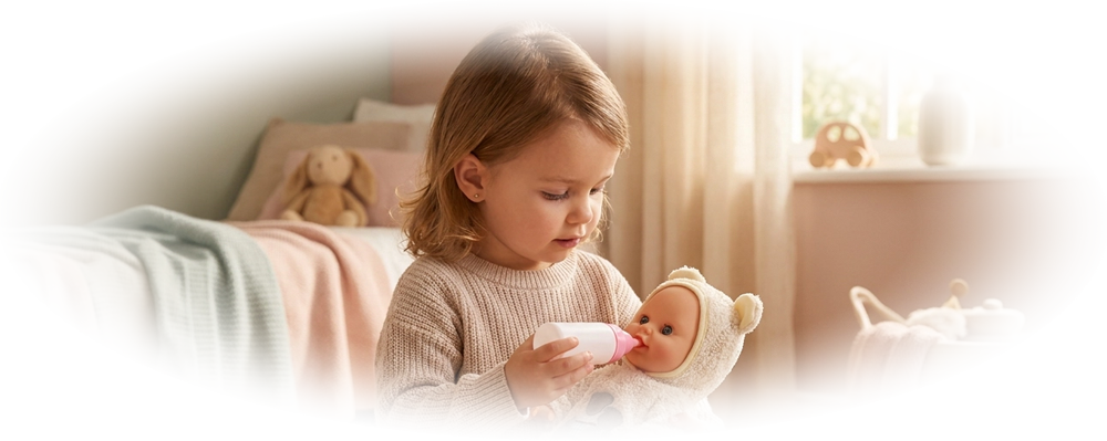 Toddler girl feeding a baby doll with a bottle while kneeling on a beige carpet in a bedroom.