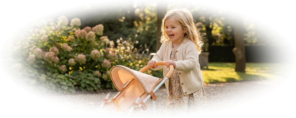 A smiling young girl pushing a beige doll stroller on green grass in a sunny garden.