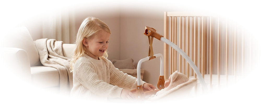 Child kneeling on a round rug, smiling and touching a baby in a beige infant swing chair.