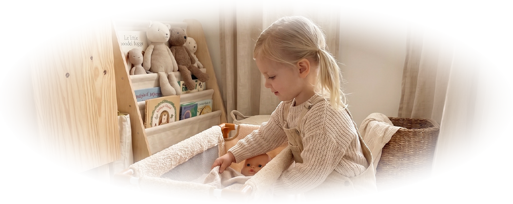 Young child kneeling beside a mesh crib, tucking a doll into a blanket in a cozy room.