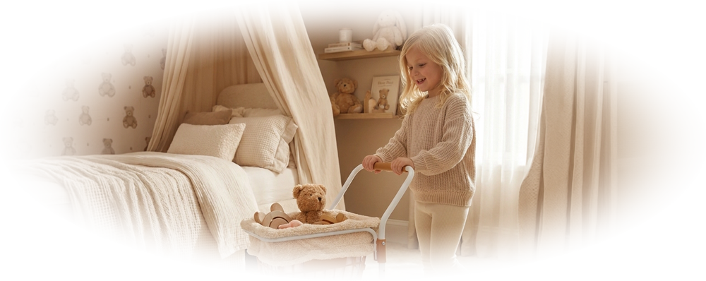Little girl in beige sweater pushing toy stroller with teddy bear in cozy bedroom.