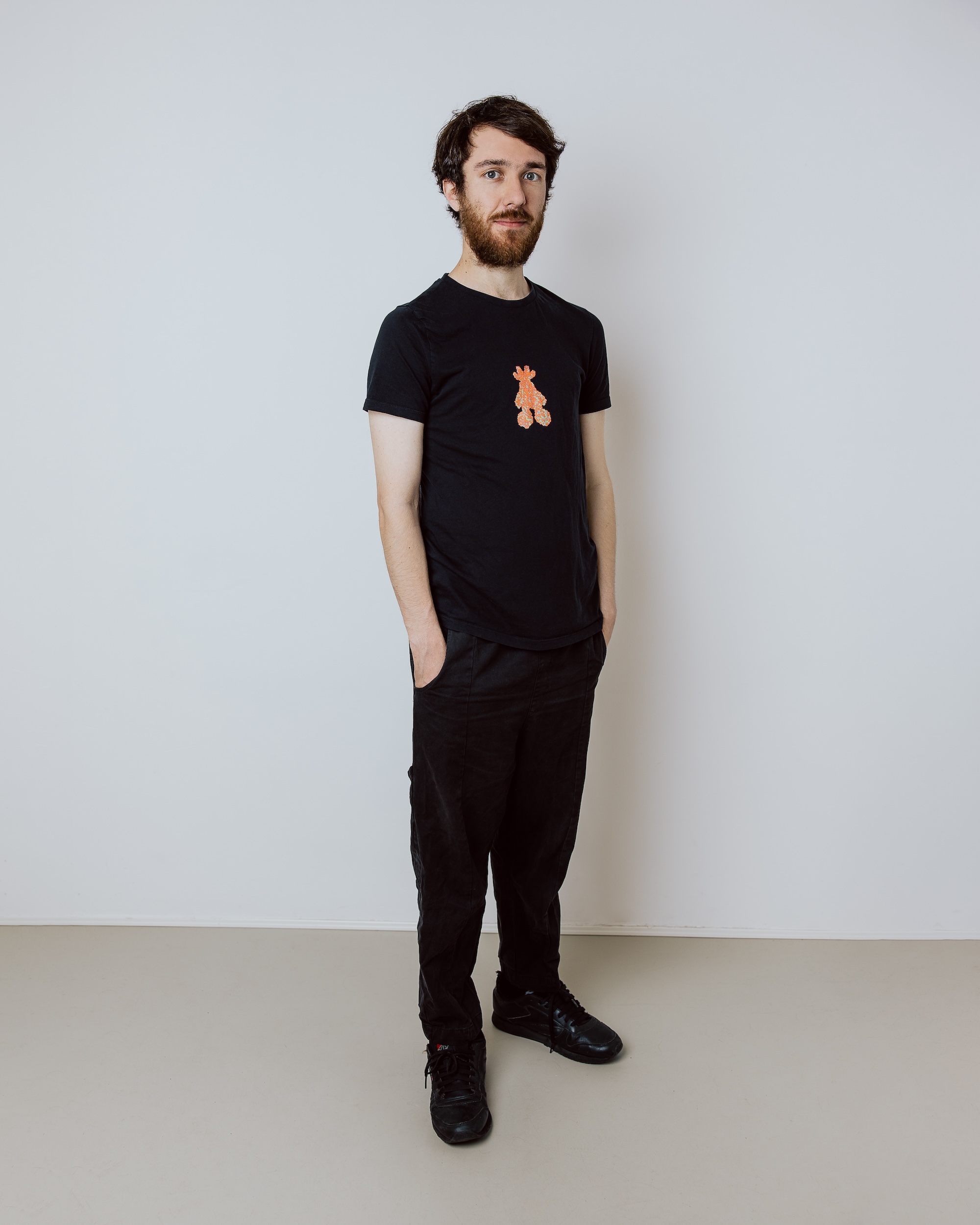 Man with beard wearing black t-shirt and pants standing against plain light wall with hands in pockets.