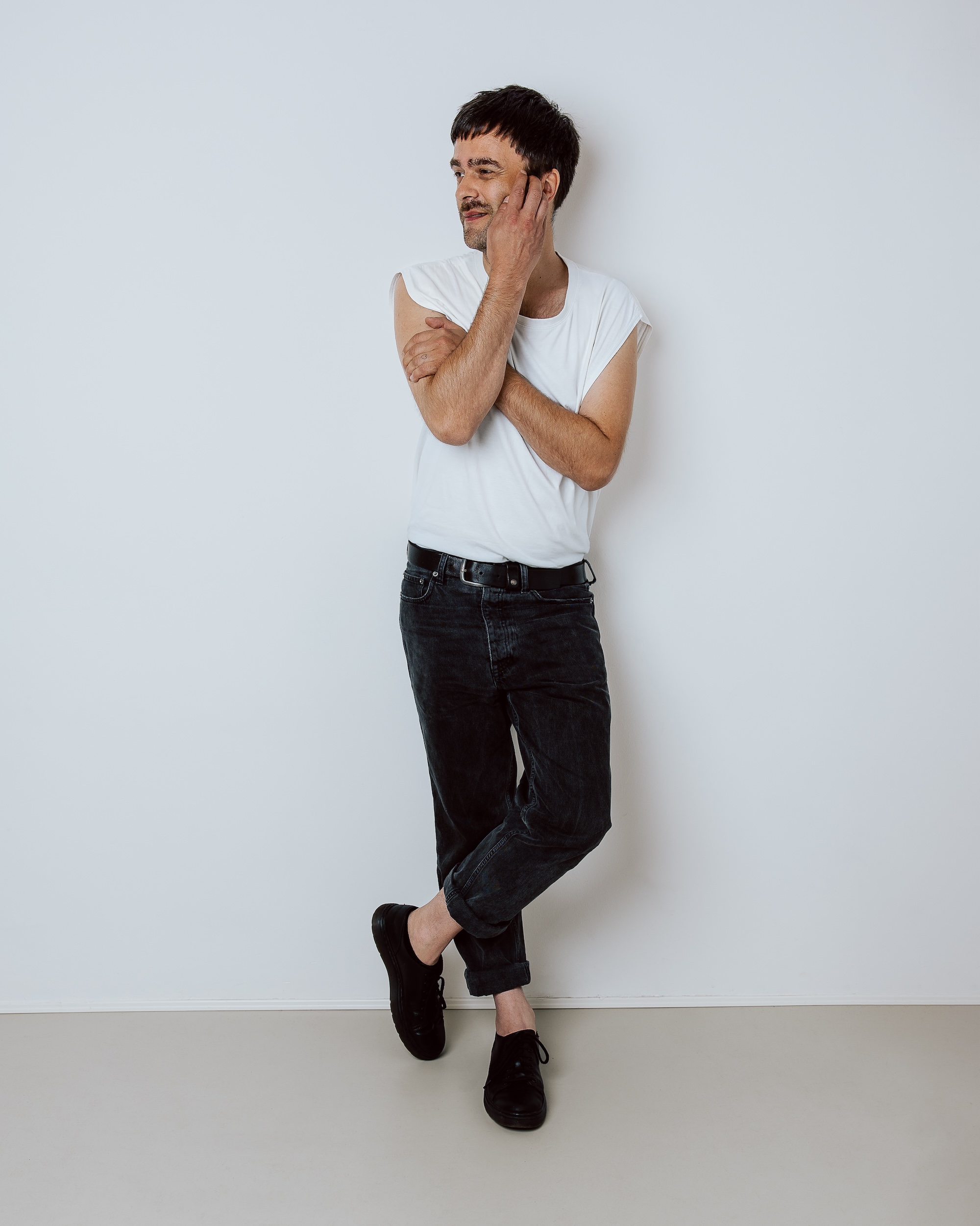 Man in white sleeveless shirt and black jeans leaning against a white wall with crossed arms.
